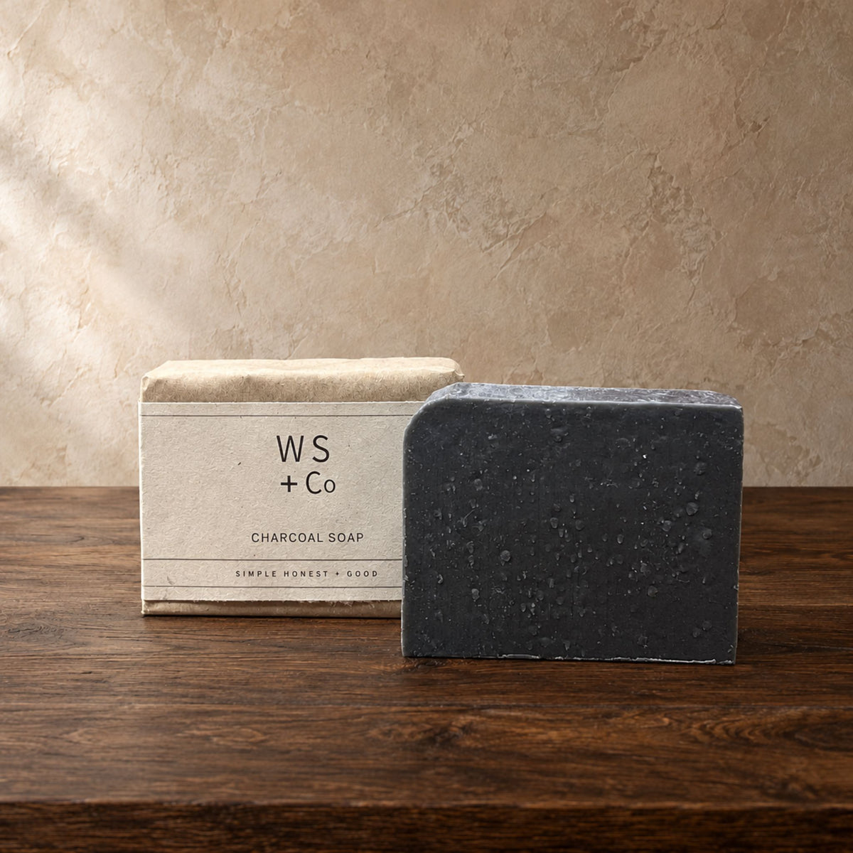 A bar of charcoal soap stands in front of its kraft paper packaging box on a dark wooden table against a textured beige wall. The box is labeled 'WS + Co CHARCOAL SOAP'.