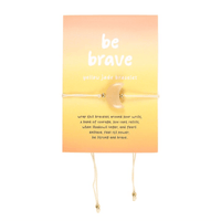 A yellow jade bracelet with a crescent moon charm displayed on a golden gradient card reading “be brave.” The adjustable cord design with small gold beads symbolises courage and strength with an uplifting message.